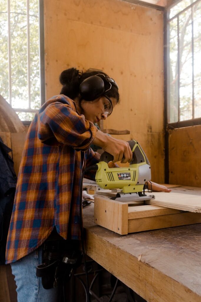 A female carpenter working with a power tool on a wooden plank in an indoor workshop setting.